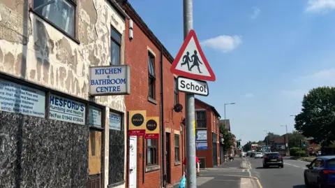 A picture of part of Chapel High Street. There is a boarded-up shop next to terrece houses. There are a number of cars on driving on the road.