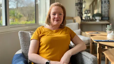 Lorna Pirozzolo is smiling at the camera with her hands folded in front of her. Wearing a bright orange T-shirt she is sitting in an armchair with a table to her left and a mirror behind her. There is window to her right through which a garden can be seen.