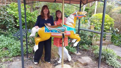 Two women stand in a garden with a brightly coloured carousel horse under a cover