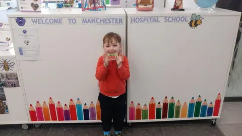 REBEKAH DALRYMPLE Hector, a young boy wearing a red jumper, he smiles brightly under a sign that reads welcome to Manchester Hospital School, with a bee sticker next to it.