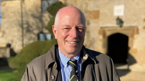 David Allard/BBC A man smiles into the camera, he is wearing a dark grey overcoat, a stripey blue shirt and a black and lime green striped tie. The sun is shining and there is a stately home entrance behind him