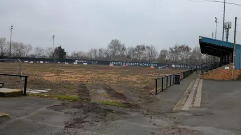 A pitch which is mostly mud, surrounded by advertising boards and lights. A spectator stand on one side of the pitch.