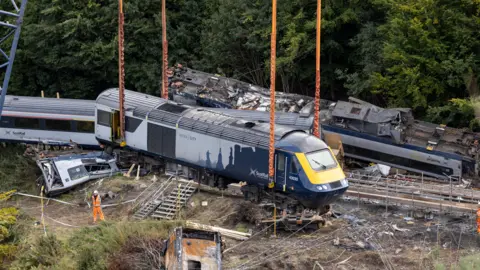 Getty Images A train carriage is raised into the air by a crane. Behind it, another carriage from the same train can be seen upside down and heavily damaged. A damaged panel from the train's frame can also be seen on the ground. Behind all this we can see a mass of thick green trees. 