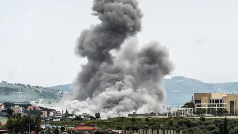 A large grey bundle of smoke rising from a village on a hillside. The smoke cloud fills most of the picture and is dramatic against the backdrop with village buildings in the foreground and hills in the background.