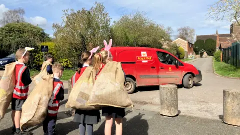 Pupils wearing postal uniform and carrying sacks of cards approach a red Royal Mail van.