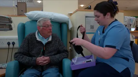 Jamie Niblock/ BBC David sits in a blue chair, whilst a nurse in blue scrubs provides a tutorial of how the bag helps him. His pouch is black all over. 