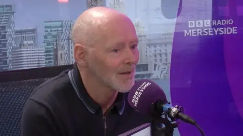 Fearon sat in the BBC Merseyside studio during his interview. The background is purple with the BBC branding. Fearon is bald and wears a dark polo zip-up.