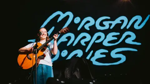Nadine Ballantyne Morgan Harper-Jones performing on stage. She has long dark hair and is wearing glasses and playing a guitar. Her name is projected on the stage behind her in blue writing.