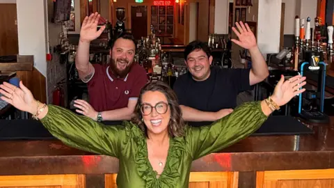 Roisin Quinn Roisin Quinn is standing in front of a wooden bar with her arms in the air. Tom Wilson and Alex Medlicott are standing behind the bar with a single arm in the air. They are all smiling at the camera. 