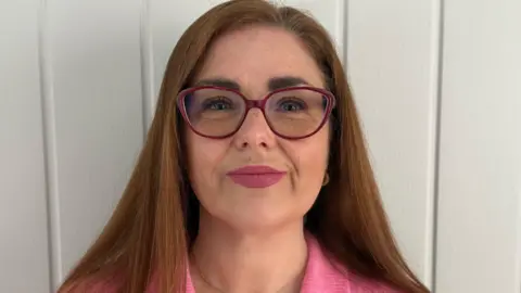 Mandy McCreight Woman with red hair and a pink shirt wearing red glasses.