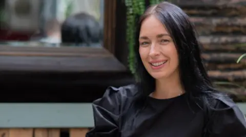 A woman wearing a black dress, with dark hair sitting on a wooden bench.