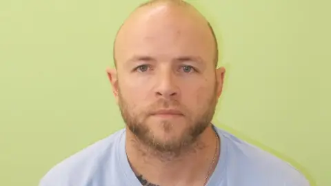 Guernsey Police Kirk Bishop with short hair and beard wearing a pale blue T-shirt against a green background