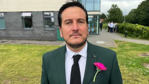 Andy Bennett. He has short dark hair and facial hair and is wearing a white shirt, black tie and green jacket. There is a pink flower pinned to his chest. He is looking directly at the camera. 
