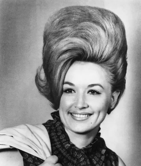 Michael Ochs Archives/Getty Images Country singer Dolly Parton poses for a portrait in 1965 in Nashville, Tennessee. In the black and white image, she has a massive beehive updo and a wide smile. A light-coloured shirt is slung over one shoulder. 