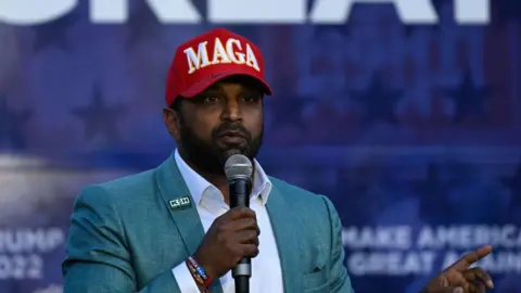 Kash Patel holds a microphone. He is wearing a green blazer and a red hat that says "MAGA" across the top. It appears to have Donald Trump's signature on the brim