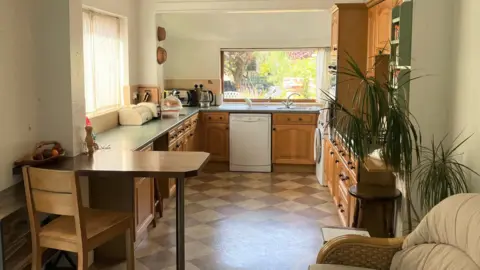 A kitchen with old-fashioned wooden cabinets and a table with one wooden chair next to it. There are two large windows, one behind the sink looks out onto a plant-filled garden.