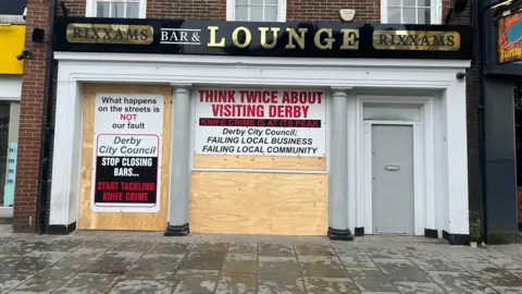 Exterior of a bar boarded up and covered in posters criticising Derby City Council about knife crime 