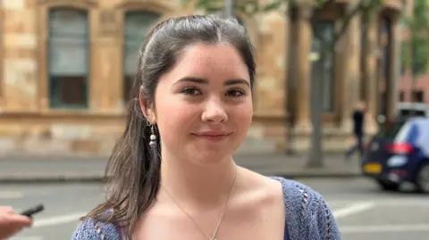 A young woman with long brown hair in a ponytail, she has brown eyes and is wearing make-up. She is wearing a blue top and has silver jewellery. The background behind her is blurred.