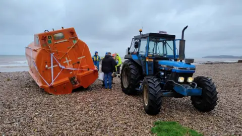 Stephen Waller A orange lifeboat being towed by a blue tractor on a pebble beach. There are coastguards between the lifeboat and tractor. The sky is cloudy and the sea in the distance. 