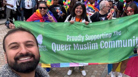 Ferhan Khan smiling in a selfie taken at a pride event with other LGBT+ Muslims in the background holding a banner