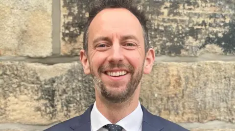 Ed Whiting smiles at the camera. He is standing against a stone wall and wearing a dark suit, white shirt and black tie with whit polka dots 
