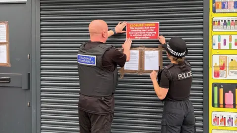 Somerset Council Two officers are putting a closure order on the front of a shop shutter. The officers are seen in uniform from the back.