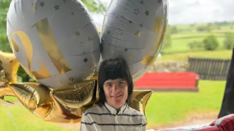Charlotte Caldwell A young man sits on a garden seat. Behind him are balloons that say 21 and in front of him is a birthday cake. The cake is Harry Potter themed with magic wand and witches hats. 