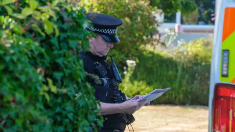 PA Media A police officer looks down at a sheert of paper while wearing uniform next to a bush and police van outside Widdecombe's home on Friday.