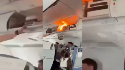 A small fire in an overhead locker inside a plane cabin. People beneath are looking up.