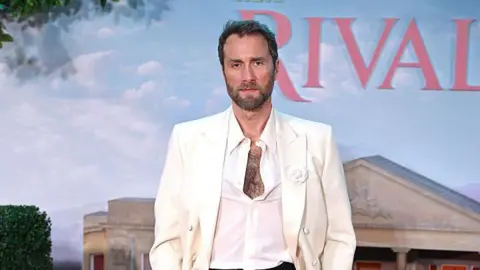 Getty Images Oliver Chris wearing a white blazer and shirt. He is standing in front of a 'Rivals' sign. 