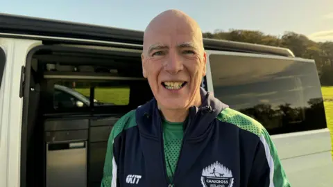 The picture features a man standing in front of a white van. He is wearing a navy and green sports jacket. Behind his van is a green field