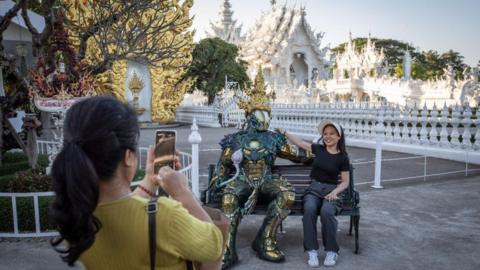 Tourists take pictures on a bench next to the White Temple. The "Wat Rong Khun" most commonly known as the "White Temple" was designed and constructed by famous Thai artist Chalermchai Kositpipat and opened to visitors in 1997.