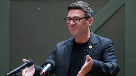 Reuters Andy Burnham stands at a podium in front of two microphones in a black suit jacket and black t-shirt.