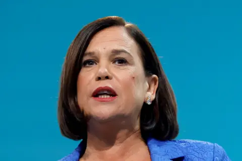 Reuters Mary Lou McDonald is speaking. She's wearing blue jacket and the backdrop is also blue.