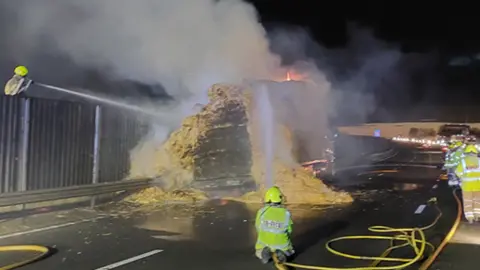 National Highways Firefighters on a motorway spraying water from hoses onto a hay lorry on fire.