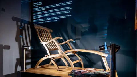 White Star Heritage A wooden deck chair is exhibited on a wooden base with a black board behind it. There is text on the board and lights shining on the chair. A tartan throw is on the base next to the chair.