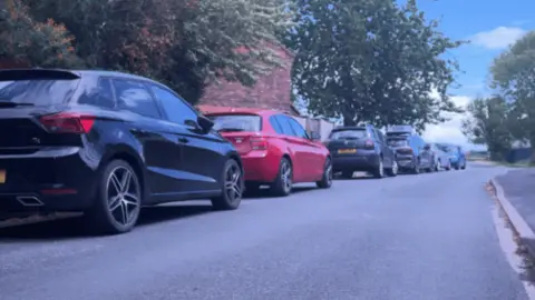 A line of cars parked on the side of a road. There is blue sky in the background and trees on the same side as the cars.