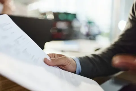 A hand holding a white piece of paper at the desk. The person is wearing a grey suit with a blue shirt beneath it. The desk behind the piece of paper is blurred.