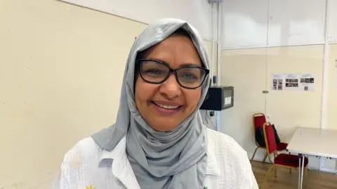 Oumaime Muhammed smiles at the camera, she wears a light grey headscarf and glasses, she is in Easton Community centre
