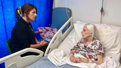 NIKKI FOX/BBC Social worker Angela Jarvis sits at the beside of Mary, a patient at Lister Hospital in Stevenage. Mary is lying in bed with a blue and white sheet over her. She is wearing a top with red flowers on a white background. Mary has white, shoulder length hair and is looking at the social worker. Her hands are clasped together. She has hospital tags on her wrist. The social worker is wearing black clothing with a clasp at the back of her hair. There is a dressing down draped over a green chair to the right of Mary.