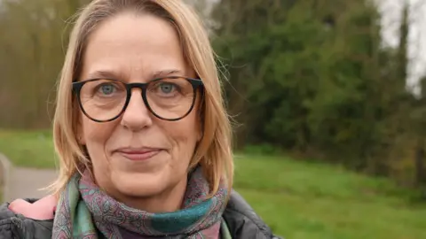 Julie Williamson standing outside by the side of the road. She has blonde hair and is wearing glasses. She is looking directly at the camera and smiling.