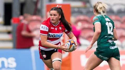 Jorja Aiona running with the ball in both of her hands held to her left during the PWR semi-final game for Gloucester-Hartpury against Trailfinders. A Trailfinders player (right) is running towards her.