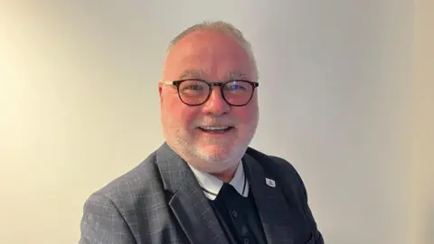 Lydia Dowling Ranera/BBC Wayne Fitzgerald headshot, smiling, wearing glasses and a check suit.