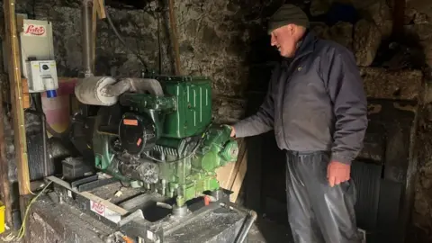 A man stands next to a green diesel generator in an old stone outbuilding. He is wearing waterproof trousers and a dirty top.