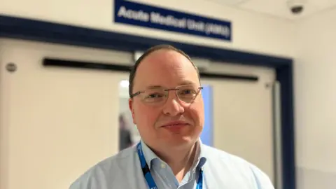 George Carden/BBC Dr Andy Heeps, chief executive of the University Hospitals Sussex NHS Foundation Trust, pictured outside the new Acute Medical Unit at the Royal Sussex County Hospital. Dr Heeps is wearing a blue shirt and blue lanyard. He is wearing glasses.