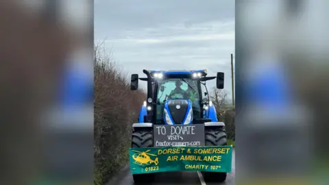 Somerset Tractor Runners A blue tractor drives down a country lane. A man is at the wheel and a woman with a small boy also sits in the cab. All are wearing high-vis jackets. On the tractor is a sign reading "Dorset and Somerset Air Ambulance Charity". A second sign reads: "To donate, visit tractor-runners.com."