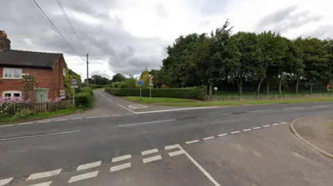 A Google street view image showing a crossroads junction. A cottage sits behind a fence on the left, behind pink flowers. A small wood is across the road on the right behind a hedge and wooden fencing. There are speed limit and other road signs by the road.