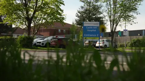 A photo taken behind grass next to a road with a blue and white sign on the other side that reads 'Castle Hill Hospital, Entrance 2'. Behind it is a small, full car park