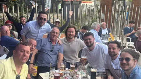 Mikey Fenton Eight men sitting around a table outside a pub. There are drinks on the table. There are also many customers sitting around them.