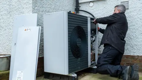 Getty Images An engineer is installing a heat pump on the side of a property. He is wearing a black jacket, trousers and boots. The building behind him is white and the large heat pump unit is visible in the foreground.